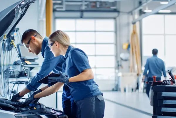 Two auto technicians in blue uniforms and safety glasses inspect a car's engine under the open hood while one holds a tablet in a bright repair shop.