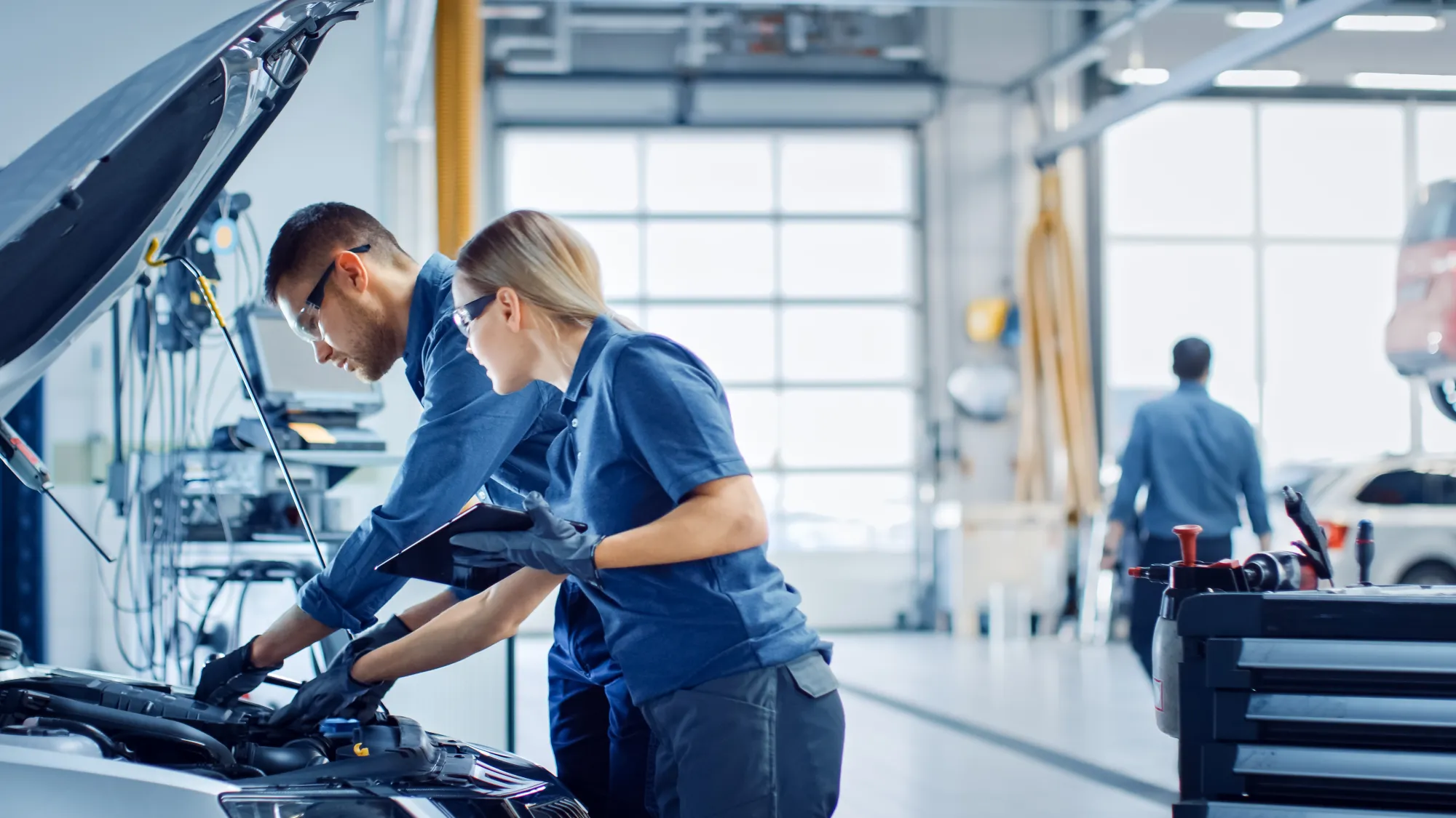 Two auto technicians in blue uniforms and safety glasses inspect a car's engine under the open hood while one holds a tablet in a bright repair shop.